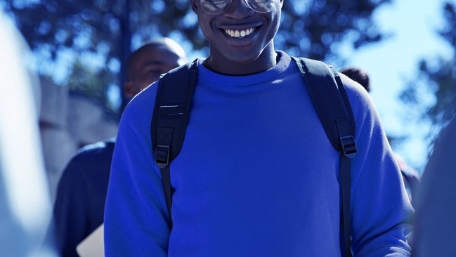 A smiling black man in a blue sweatshirt with a backpack on, clearly is walking to class.