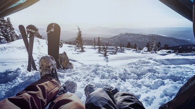 Point of view shot from people lying down in a tent looking out over a snowy mountain, there are a pair of skis and a snowboard seen to the left.